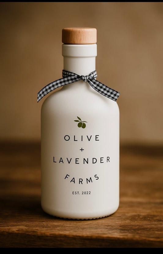 White bottle with wooden cap and checkered ribbon on a wooden surface, featuring 'Olive + Lavender Farms' branding.