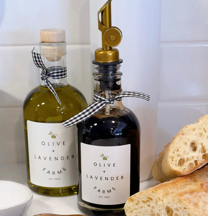 One bottle of dark balsamic vinegar and one of olive oil from Olive & Lavender Farms on a kitchen counter with bread.