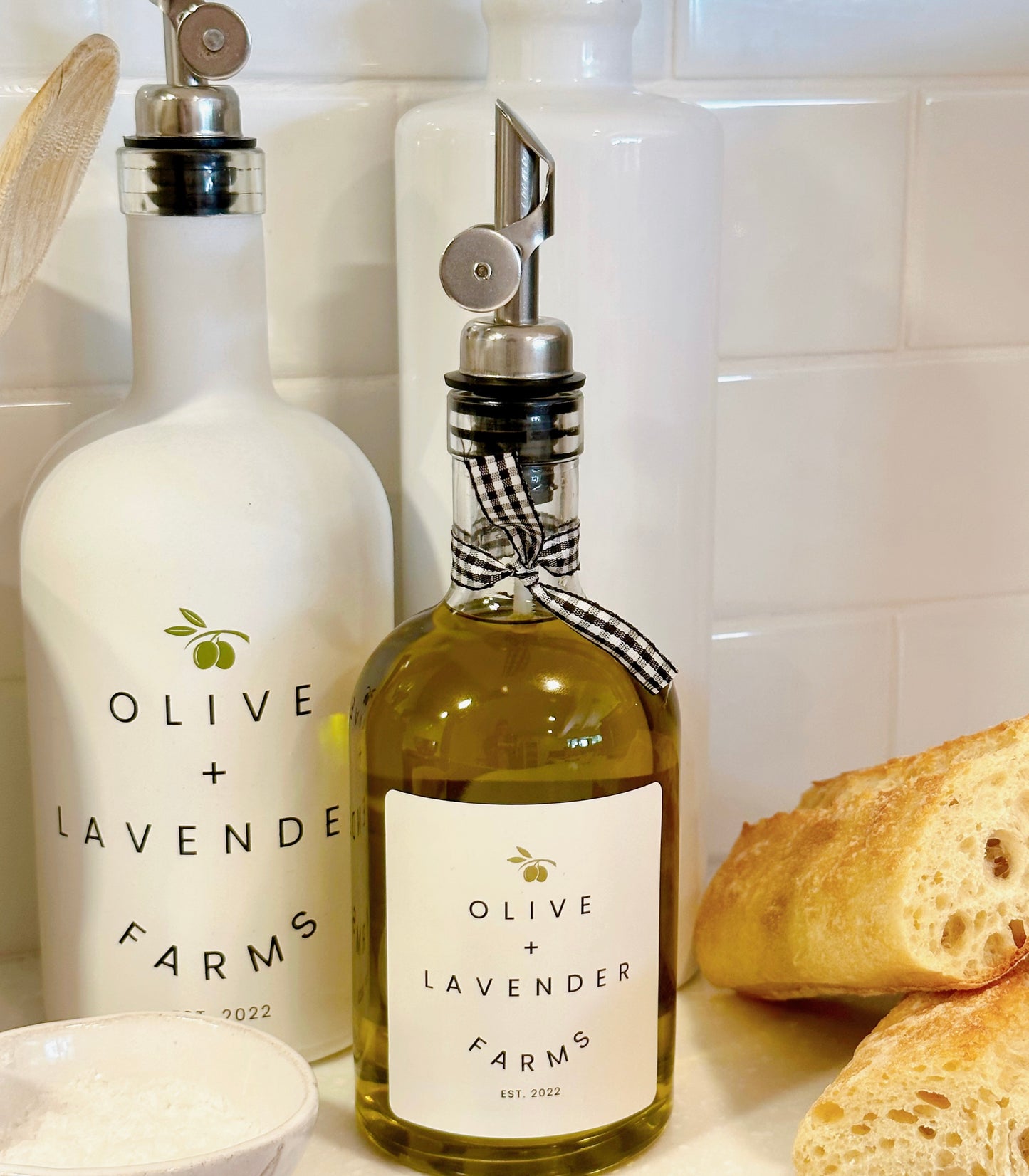 Two bottles of olive oil from Olive + Lavender Farms on a kitchen counter with bread in the background.