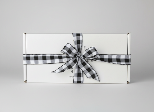 White gift box with a black and white checkered ribbon on a woven surface with greenery in the background